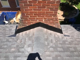 Dark metal V-shaped flashing at roof peak meeting brick chimney with asphalt shingles